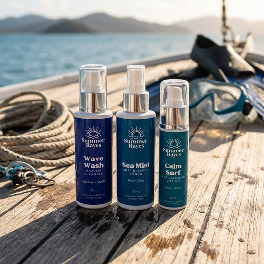 Three Summer Rayes skincare bottles (Wave Wash Cleanser, Sea Mist Toner, and Calm Surf Lotion) arranged on a rustic wooden dock with a blurred background of water, rope, and diving gear.