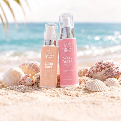 Two Summer Rayes skincare products, Calm Surf Lotion and Wave Wash Cleanser, standing on a sandy beach with seashells and a blurred ocean background.