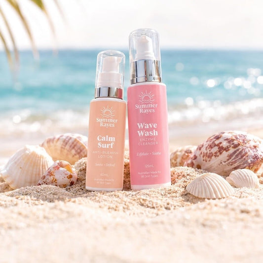 Two Summer Rayes skincare products, Calm Surf Lotion and Wave Wash Cleanser, standing on a sandy beach with seashells and a blurred ocean background.