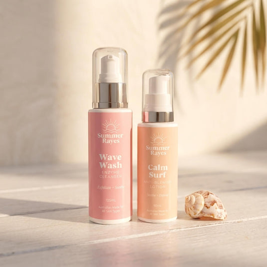 Two Summer Rayes skin care bottles, Wave Wash Cleanser in pink and Calm Surf Moisturizer in peach, standing on a light wooden surface with a seashell and palm leaf shadows.
