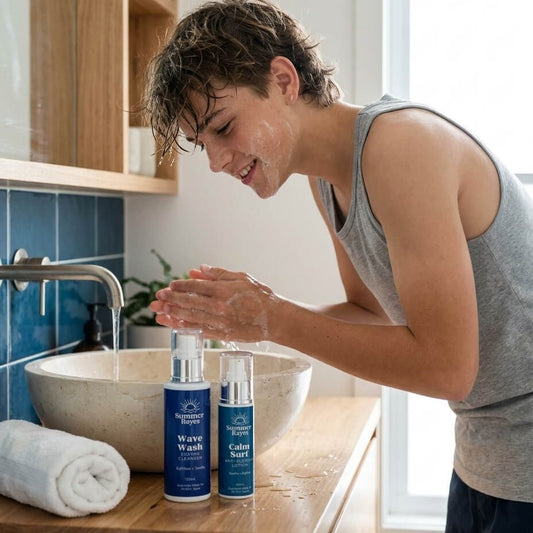 A teen boy stands at a bathroom sink, washing his face. Two blue skincare bottles from the brand 'Summer Rayes' are placed on the wooden counter in front of him.