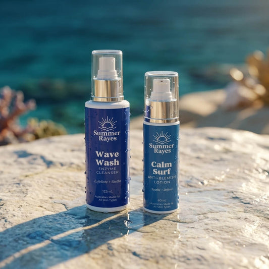 Two Summer Rayes skin care bottles, Wave Wash Enzyme Cleaner and Calm Surf Anti-Blemish Lotion, standing on a wet stone surface with a blurred ocean background.