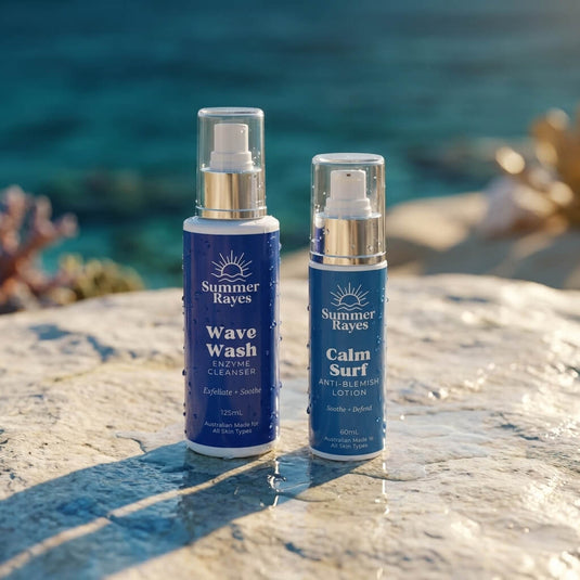 Two Summer Rayes skin care bottles, Wave Wash Enzyme Cleaner and Calm Surf Anti-Blemish Lotion, standing on a wet stone surface with a blurred ocean background.