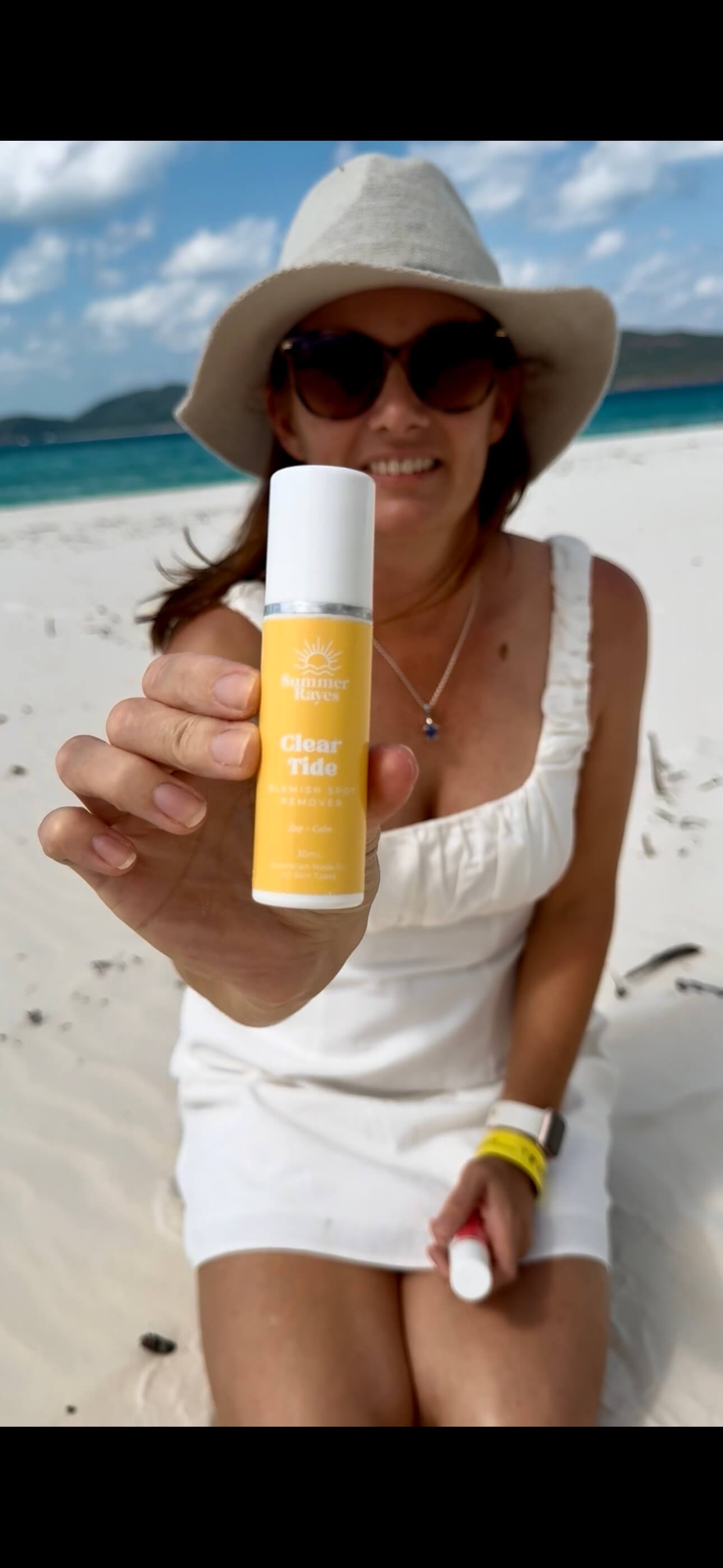 A mum of teens sitting on the beach holding out a bottle of Summer Rayes skincare