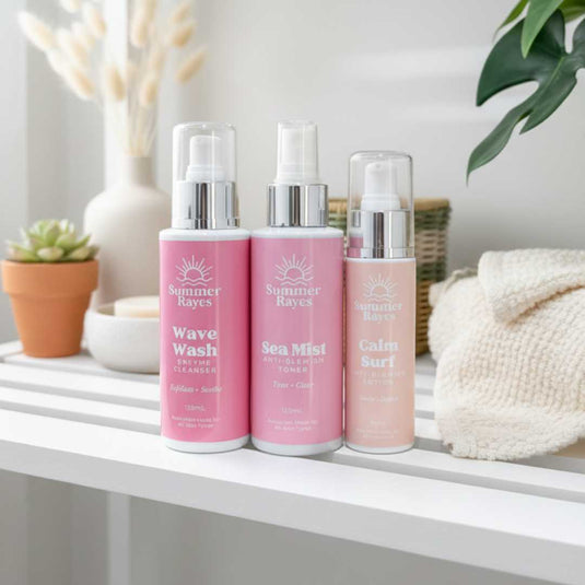 Three pink bottles of Summer Rayes products on a white surface with decorative items in the background.